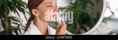 Porträt einer jungen Frau mit schönem Gesicht Anwendung Lippenbalsam, stehend im Badezimmer mit Pflanzen geschmückt Stockfoto