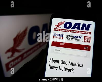 Person mit Mobiltelefon und Website des US-Medienunternehmens One America News Network (OANN) auf dem Bildschirm mit Logo. Konzentrieren Sie sich auf die Mitte des Telefondisplays. Stockfoto