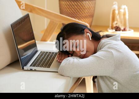 Müde schlafende Studentin mit Ohrstöpsel im Ohr, die mit geöffnetem Laptop auf dem Schreibtisch gelehnt ist Stockfoto