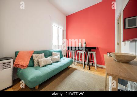 Wohnzimmer einer Kurzzeitwohnung mit einem grünen Schlafsofa, einer rot gestrichenen Wand und einem Anrichte aus Eichenholz und einem schwarzen Tisch Stockfoto