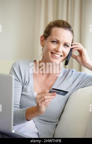 Alles, was ich will, steht mir zur Verfügung. Eine junge Frau, die auf ihrem Handy spricht, während sie eine Bankkarte in der Hand hält und mit einem Laptop sitzt. Stockfoto