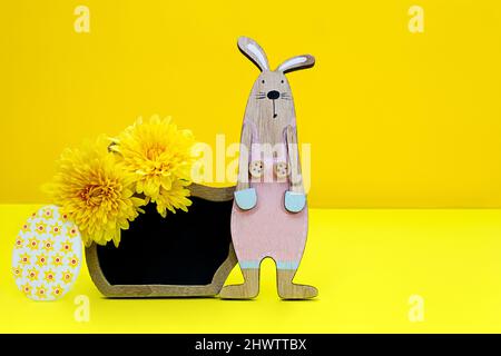 Holzkaninchen mit schwarzem Brett für Text und Chrysanthemen, ein Ei auf gelbem Hintergrund. Osterkonzept. Speicherplatz kopieren Stockfoto