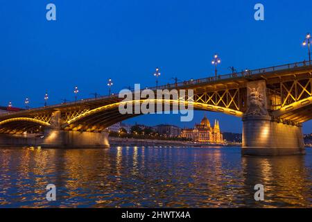 Die Margaretenbrücke mit dem Parlamentsgebäude im Hintergrund in Budapest, Ungarn, beleuchtet über der Donau bei Nacht. Stockfoto