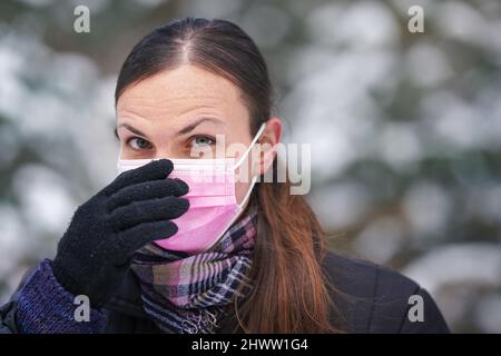 Junge Frau in warmer Winterkleidung, Schutzhandschuhe tragen, Einweg-Einweg-rosa Gesicht Virus Mundmaske setzen oder entfernen, Nahaufnahme, verschwommen Stockfoto