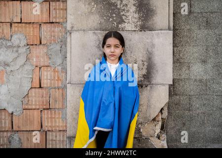 Kleines Mädchen mit ukrainischer Flagge vor einer durch Bomben zerstörten Mauer. Das kleine Mädchen schwenkt die Nationalflagge und betet für den Frieden Stockfoto