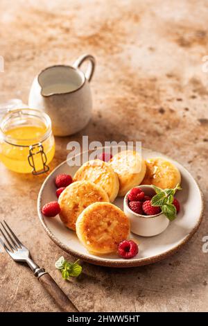 Hüttenkäse-Pfannkuchen, Syrniki mit Honig und frischer Himbeere auf Keramikplatte auf texturiertem Hintergrund Stockfoto