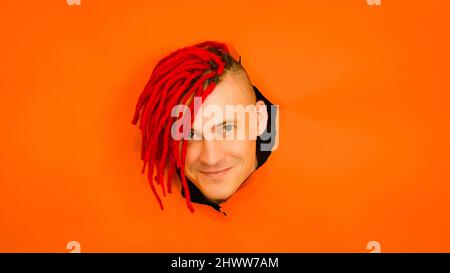 Fröhlicher junger Mann mit gefärbtem Haar, der im Studio im Loch auf orangefarbenem Hintergrund auf die Kamera schaut Stockfoto