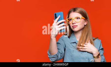 Fokussiertes Mädchen in einem einfachen blauen Hemd und einer orangefarbenen Brille, das auf das Handy schaut. Frau nutzt Handy, scrollt durch soziale Netzwerke, sucht nach tollen Angeboten, findet die neuesten Nachrichten heraus Stockfoto