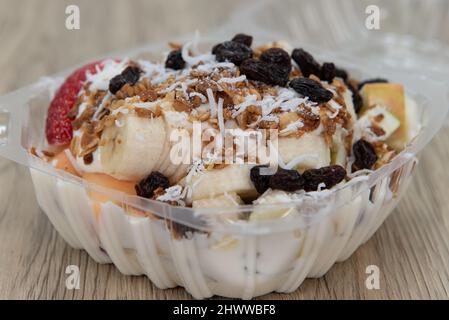 Köstliche und gesunde Obstschale mit Banane, Rosinen, Kokosnuss und Joghurt, die alle in einem Take-Out-Karton serviert werden. Stockfoto