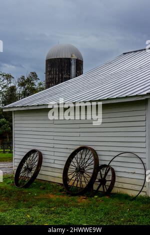 Alte, rostige Wagenräder lehnen sich an ein weißes Farmgebäude mit einem Silo im Hintergrund. Stockfoto