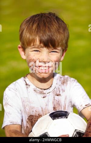 Spielen möchten. Porträt eines schlammigen kleinen Jungen, der einen Fußball hält. Stockfoto