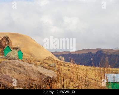 Hoch in den vergilbten Herbstbergen des Kaukasus unter bewölktem Himmel stehen unter den Felsbrocken grüne temporäre Holzhütten. Stockfoto