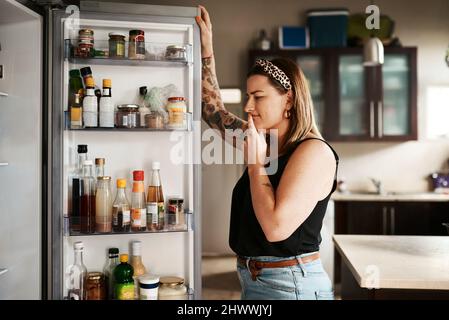 Hmm... was man zum Abendessen machen soll. Aufnahme einer jungen Frau, die zu Hause in einem Kühlschrank sucht. Stockfoto