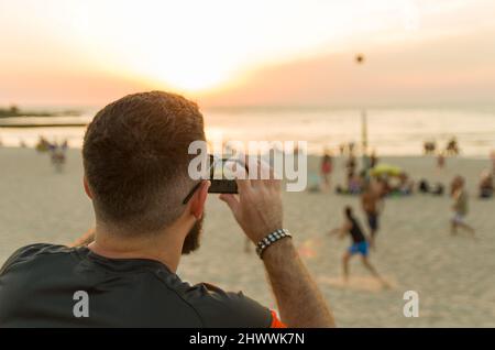 Mann fotografiert den Sonnenuntergang am Strand mit dem Smartphone Stockfoto