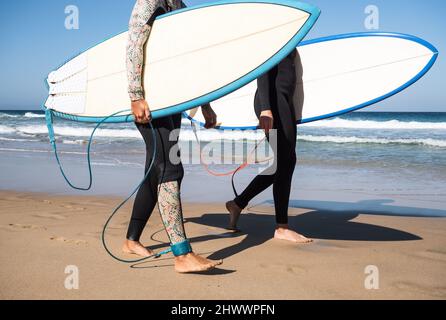 Nicht erkennbare Surfer-Weibchen, die am Strand mit Surfbrettern und Winter-Neoprenanzügen spazieren Stockfoto