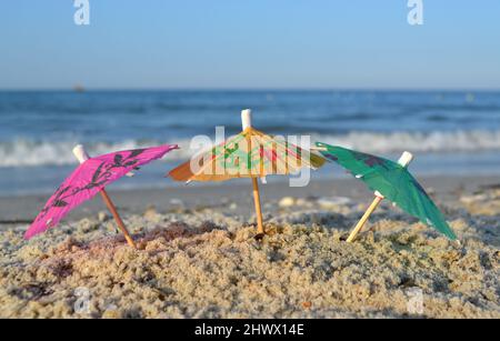 Drei kleine Papier-Cocktail-Schirme stehen im Sand am Sandstrand aus nächster Nähe Stockfoto