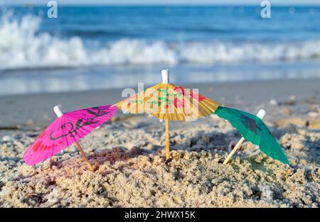 Drei kleine Papier-Cocktail-Schirme stehen im Sand am Sandstrand aus nächster Nähe. Stockfoto