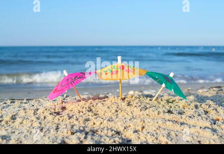 Drei kleine Papier-Cocktail-Schirme stehen im Sand am Sandstrand aus nächster Nähe. Stockfoto