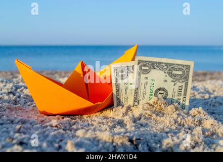 Orangefarbenes Papierboot und zwei Papierdollar-Scheine, die zur Hälfte im Sand am Sandstrand vergraben sind Stockfoto