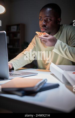 Afrikanischer Geschäftsmann, der Pizza isst und an seinem Arbeitsplatz sitzt und während der Frist im Büro online am Laptop arbeitet Stockfoto