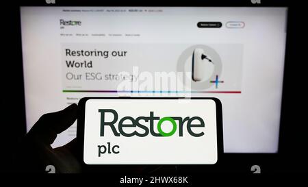 Person, die Mobiltelefon mit dem Logo des britischen Datenverwaltungsunternehmens Restore plc auf dem Bildschirm vor der Business-Webseite hält. Konzentrieren Sie sich auf die Telefonanzeige. Stockfoto