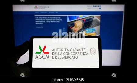 Person, die Mobiltelefon mit dem Logo der italienischen Wettbewerbsbehörde (AGCM) auf dem Bildschirm vor der Unternehmenswebseite hält. Konzentrieren Sie sich auf die Telefonanzeige. Stockfoto