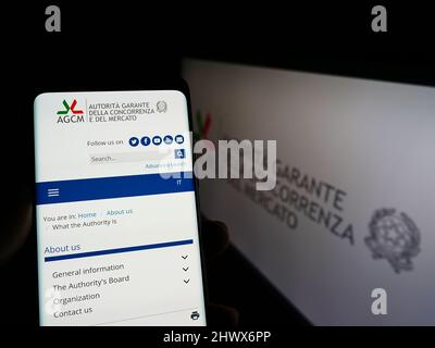 Person, die Mobiltelefon mit Weboage der italienischen Wettbewerbsbehörde (AGCM) auf dem Bildschirm vor dem Logo hält. Konzentrieren Sie sich auf die Mitte des Telefondisplays. Stockfoto
