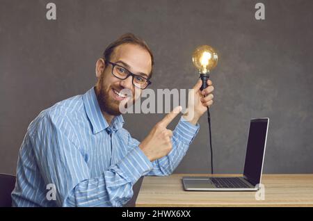 Junger, glücklicher Geschäftsmann, freiberuflicher Mitarbeiter mit Glühbirne vorne am Laptop am Schreibtisch Stockfoto