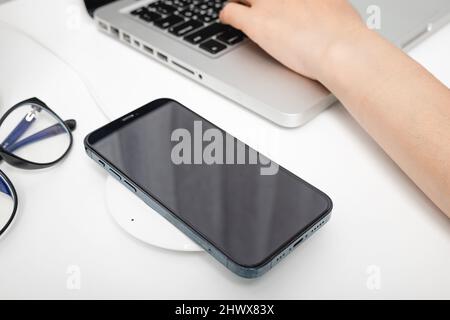 Aufladen des Smartphone-Akkus mit einem kabellosen Ladegerät in der Nähe des Laptops. Moderne Technologie überträgt Energie-Mobiltelefone auf den Desktop am Arbeitsplatz bei Stockfoto