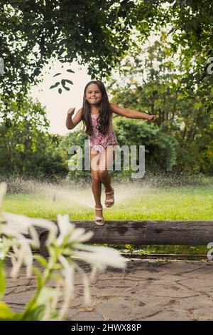 Junge strahlende lächelnde Mädchen, die Spaß haben, aus dem Baumstamm zu springen, sich zu verwöhnen und draußen im Park mit Bäumen und Sträuchern im Hintergrund tagsüber zu spielen. Träumen Stockfoto