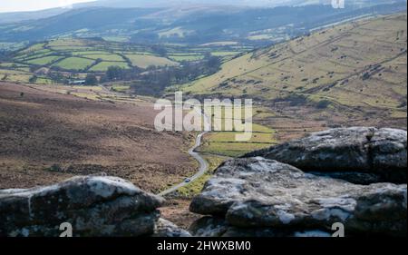 Eine hell erleuchtete, kurvenreiche Straße schlängelt sich durch den Dartmoor-Nationalpark. Ein weicher Vordergrund verstärkt das Maßstabsgefühl, wenn die Straße durch ein Tal führt Stockfoto