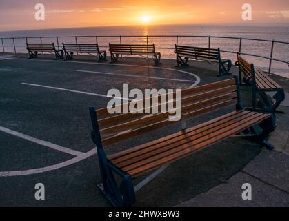 Eine geschwungene Reihe von sechs Holzbänken zeigt an einem kalten, klaren Morgen auf das Meer. Bernsteinfarbenes Sonnenlicht bringt sie in ein orangefarbenes Leuchten, während sie leer stehen Stockfoto