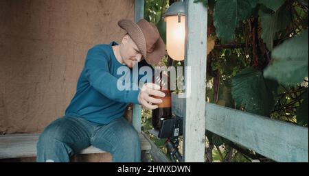 Cowboy-Mann auf der Veranda trinkt Bier aus einer Flasche und kommuniziert online per Telefon Stockfoto