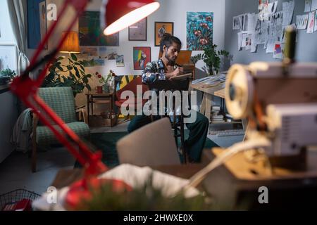 Moderner junger Mann in modischer Freizeitkleidung, sitzend auf einem Stuhl im Atelier der Künste oder in einer Werkstatt und ziehend auf einem Notizblock Stockfoto