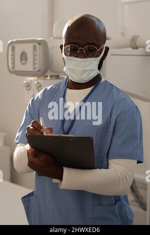 Junger seriöser afroamerikanischer Arzt in Uniform und Schutzmaske, der Notizen oder Rezepte für den Patienten im Krankenhaus macht Stockfoto