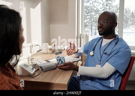 Junger afroamerikanischer Arzt in blauer Uniform hält Glas mit Pillen und empfehlen es weiblichen Patienten im Krankenhaus Stockfoto