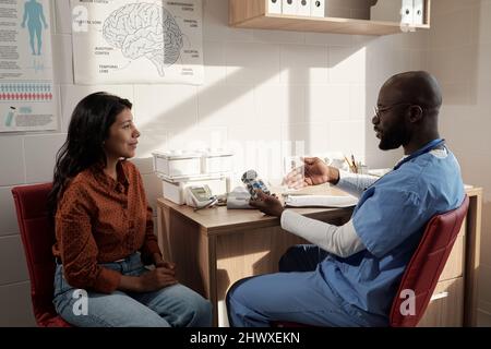 Ein junger, selbstbewusster Arzt in Uniform empfiehlt jungen hispanischen weiblichen Patienten neue Pillen und beschreibt ihre Eigenschaften Stockfoto