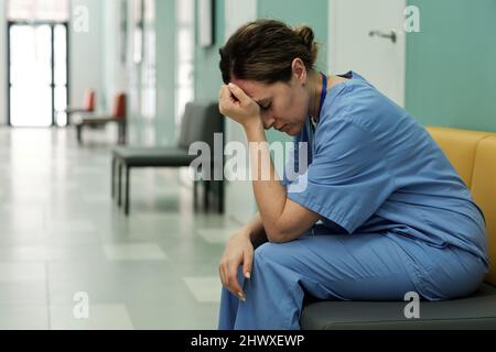 Junge müde oder gestresste Ärztin in Uniform sitzt im Krankenhausflur mit dem Kopf in den Händen nach schockierenden Nachrichten Stockfoto