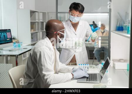 Junge asiatische Wissenschaftlerin in Schutzmaske und Laborkittel, die ihren afroamerikanischen männlichen Kollegen mit blauer Flüssigkeit zeigt Stockfoto