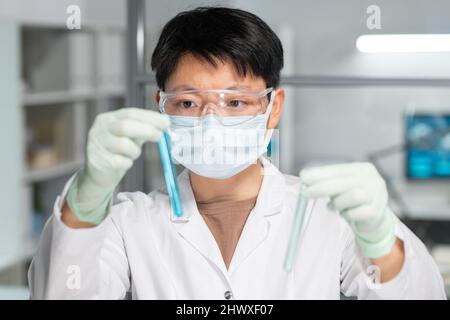 Asiatische Wissenschaftlerin in Schutzbrille, Maske und Handschuhen, die zwei Proben von flüssigen Substanzen vergleicht Stockfoto