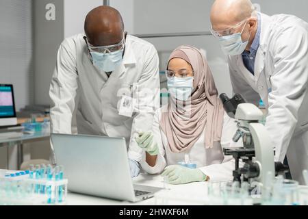 Junge muslimische Ärztin zeigt auf den Laptop-Bildschirm, während sie zwei männlichen Kollegen die Ergebnisse eines wissenschaftlichen Experiments erklärt Stockfoto