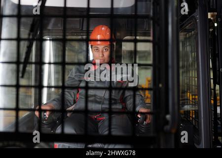 Arbeiterin in der Fabrik in Hardhat und Arbeitskleidung, die durch Gangwechsel gehalten wird, während sie die Baumaschine fährt und nach vorne schaut Stockfoto