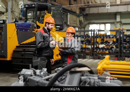 Zwei junge Arbeiter in Industrieanlagen diskutieren technische Daten in einem Dokument, das von einer afroamerikanischen Ingenieurin gehalten wurde Stockfoto