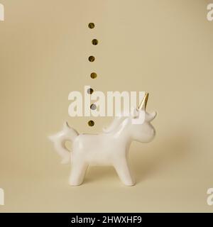 Weißes Einhorn-Sparschwein mit Goldmünzen auf minimalem gelben Hintergrund Stockfoto