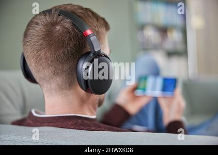Rückansicht Des Mannes, Der Sich Auf Dem Sofa Entspannt, Mit Kabellosen Kopfhörern, Die Filmmusik Oder Podcast Vom Mobiltelefon Streamen Stockfoto