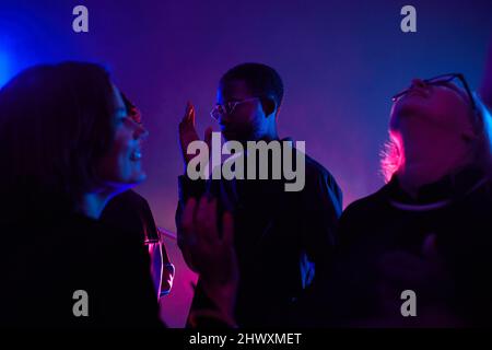 Hinterleuchtetes Porträt eines schwarzen jungen Mannes, der in Menschenmengen tanzt, während er in einem rauchigen Club, der von Neon beleuchtet ist, in einem Kopierraum feiert Stockfoto