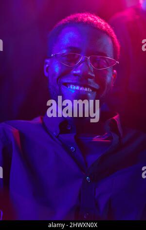 Vertikales Porträt eines jungen afroamerikanischen Mannes, der in einem von Neonlichtern beleuchteten Club lächelt Stockfoto