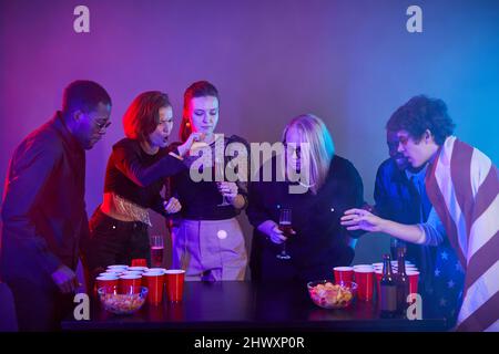 Seitenansicht verschiedene Gruppe von Menschen spielen Trinkspiele auf Party in Neonlicht und Spaß haben Stockfoto