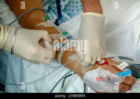 Die Hand eines Krankenpflegers mit Handschuhen injiziert Flüssigkeit in einen Katheter, der am Arm eines Patienten befestigt ist. Stockfoto