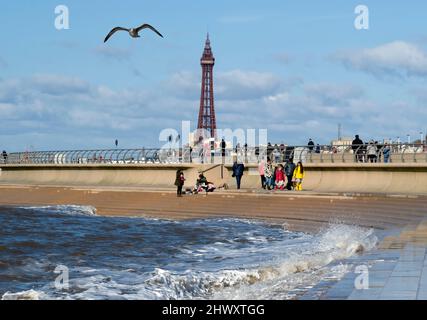 An einem hellen, sonnigen Tag treffen Urlauber auf den Sandstrand im Schatten des weltberühmten Blackpool Tower in Blackpool, Lancashire, Großbritannien Stockfoto